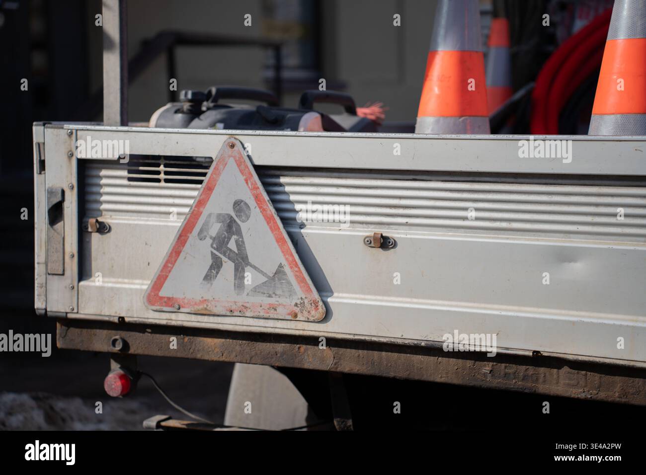 portellone per camion da lavoro urbano illuminato dal sole, con un cartello stradale triangolare con agenti atmosferici, coni stradali arancioni e cavi a spirale; pannelli metallici usurati, sporcizia a. Foto Stock