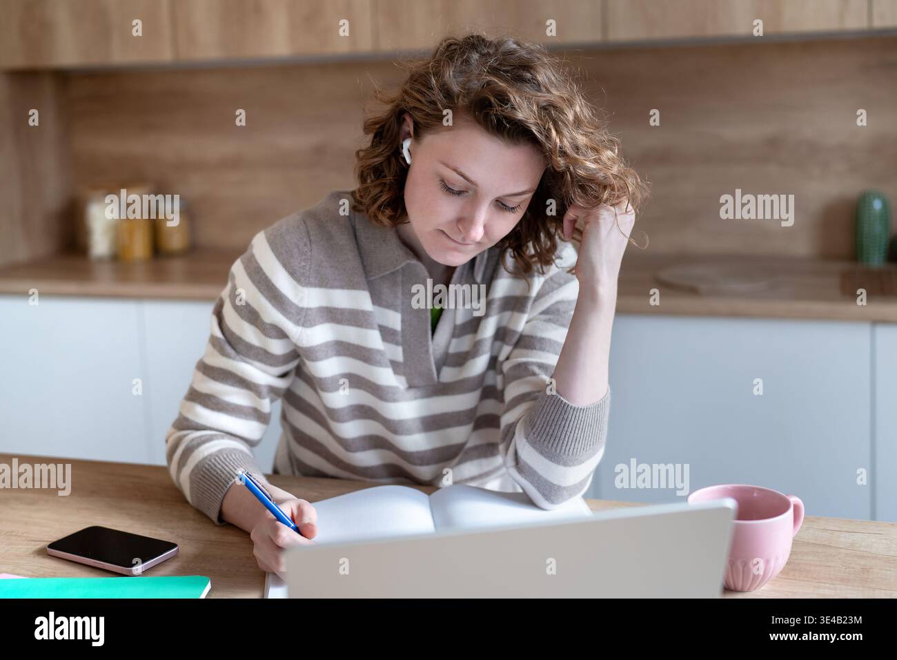 Studentessa entusiasta che studia a casa scrivendo appunti, ascoltando il webinar online indossando gli auricolari. Foto Stock