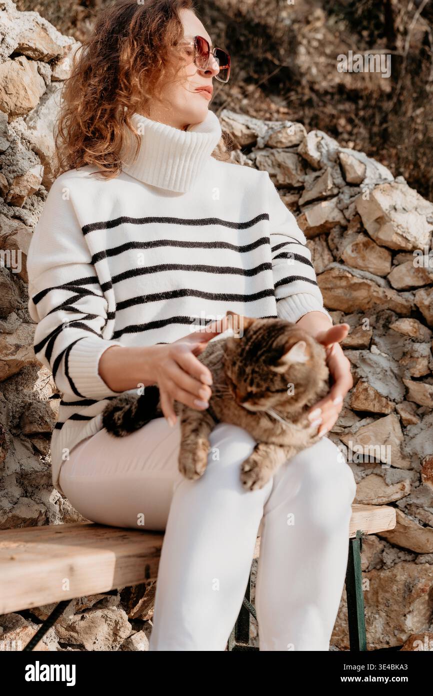 Gatto donna che siede su una panchina rustica mentre indossa un maglione a righe e coccola la sua compagna di tavolo vicino al muro di pietra Foto Stock