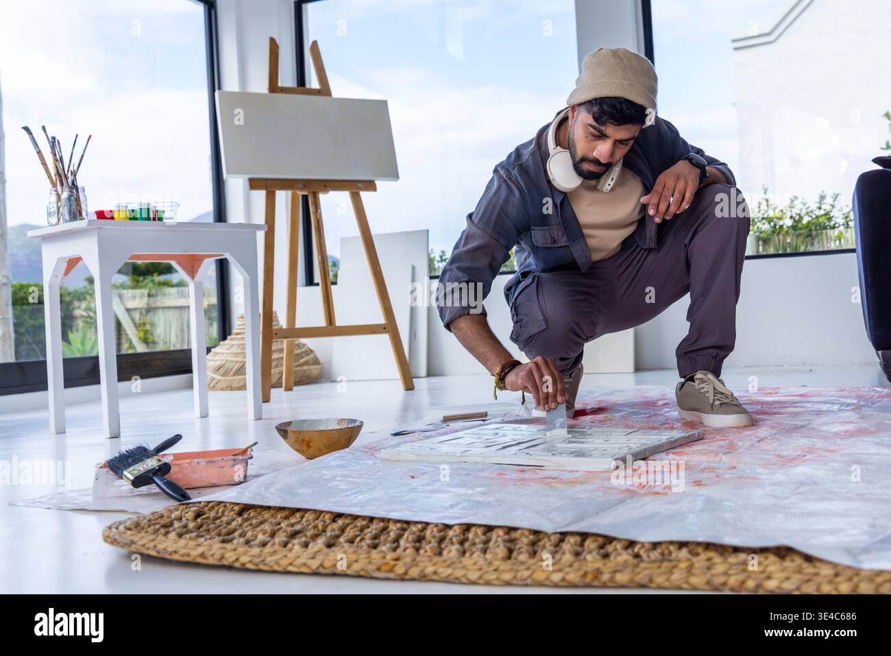 Artista maschile con barba inginocchiato che raschia la vernice su tela sul pavimento dello studio con cavalletto, vasetti, spazio copia Foto Stock