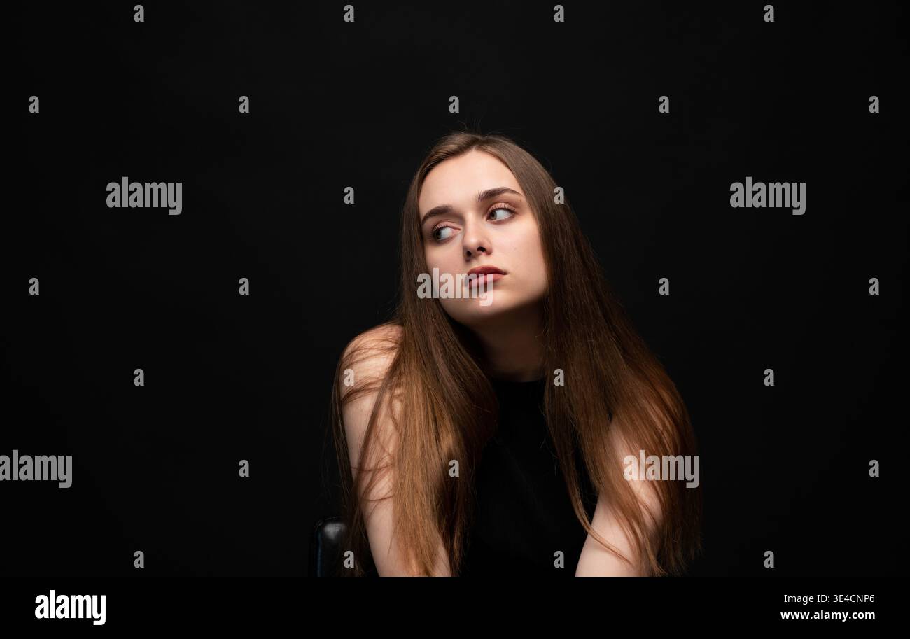 Giovane donna con lunghi capelli marroni che posa, guarda lontano, riflette, contemplando pensieri su sfondo nero Foto Stock