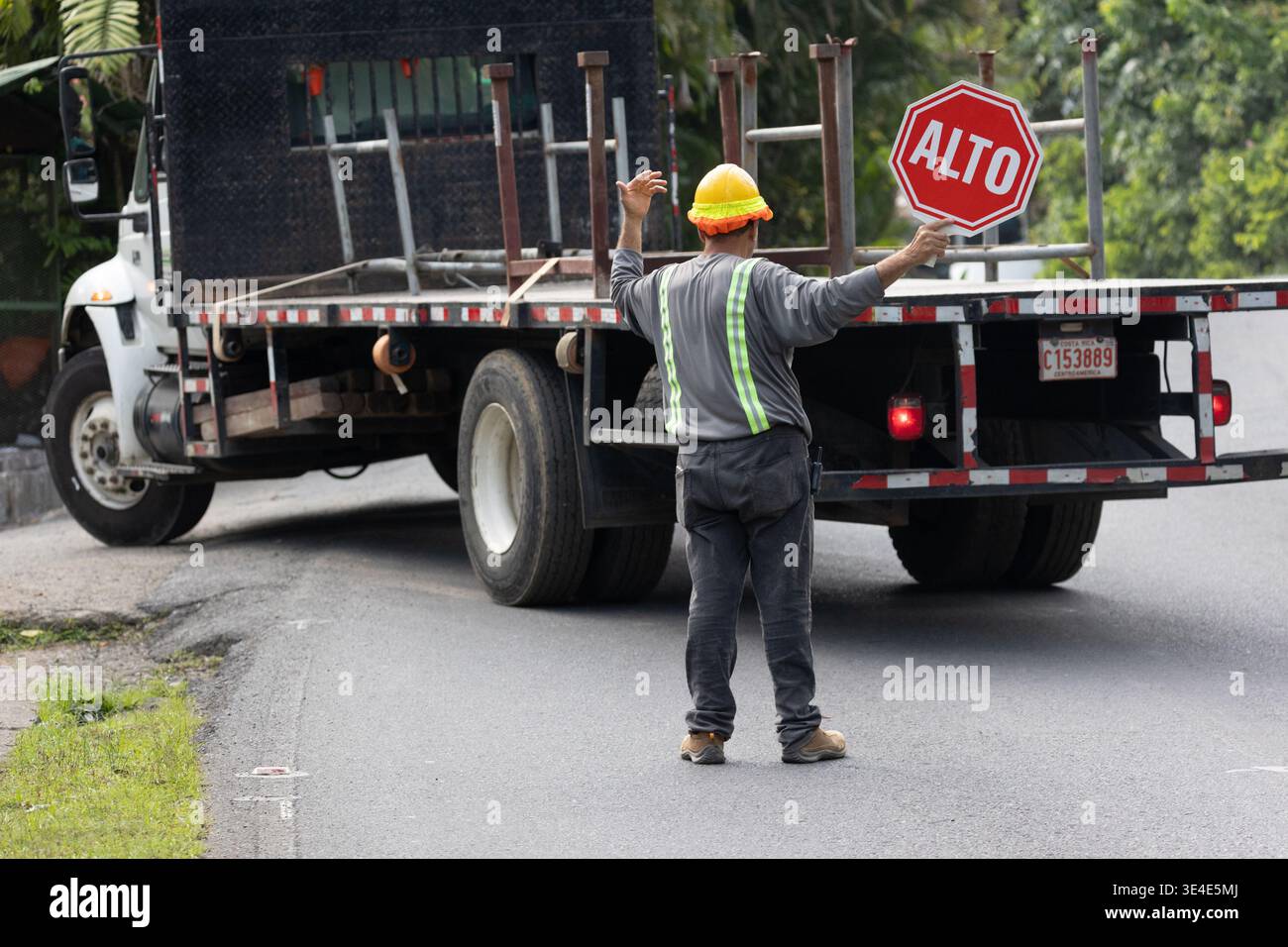 Manuel Antonio, Costa Rica - 9 agosto 2025: Un lavoro su strada con casco di sicurezza regge un segnale DI STOP "ALTO" mentre un camion procede in retromarcia su una strada Foto Stock