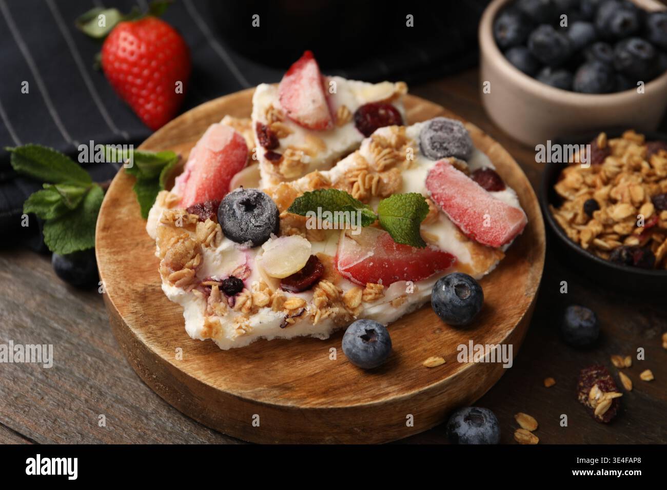 Pezzi di gustosa corteccia di yogurt con frutti di bosco, menta e muesli su tavola di legno, primo piano Foto Stock