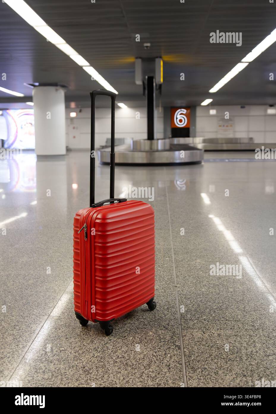 Valigia rossa in piedi nell'area di ritiro bagagli dell'aeroporto vicino alla giostra con il numero di targa nell'interno del terminal moderno. Foto Stock