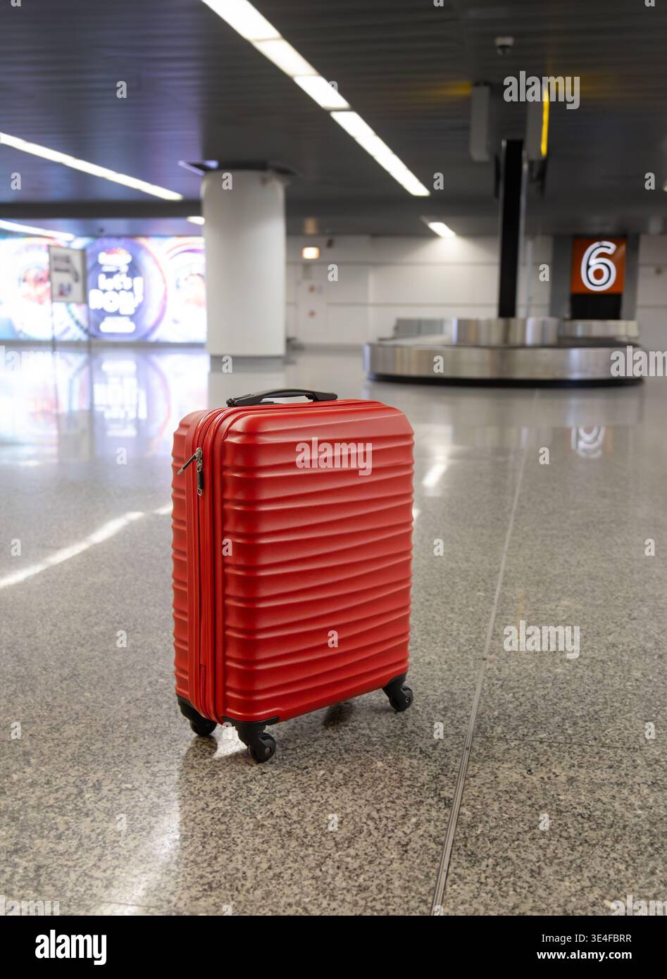 Valigia rossa in piedi nell'area di ritiro bagagli dell'aeroporto vicino alla giostra con il numero di targa nell'interno del terminal moderno. Foto Stock