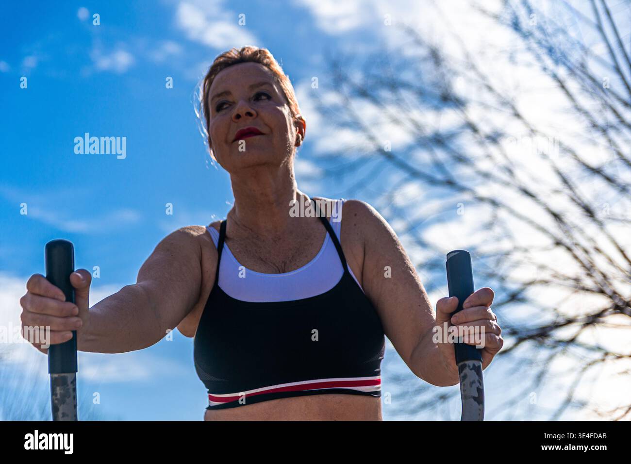 Donna anziana impegnata attivamente in un allenamento all'aperto, eseguendo esercizi cardio su una macchina per il fitness pubblica sotto un cielo azzurro limpido, promuovendo una salute Foto Stock