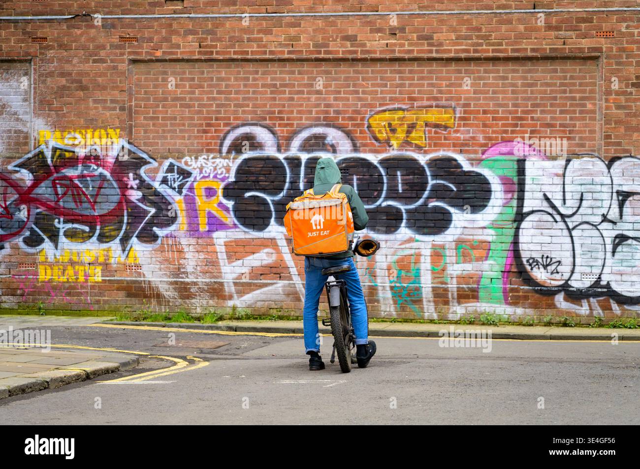 Un ciclista che consegna il cibo appena mangiato stazionario su una strada della città con graffiti su un muro, nel Regno Unito Foto Stock