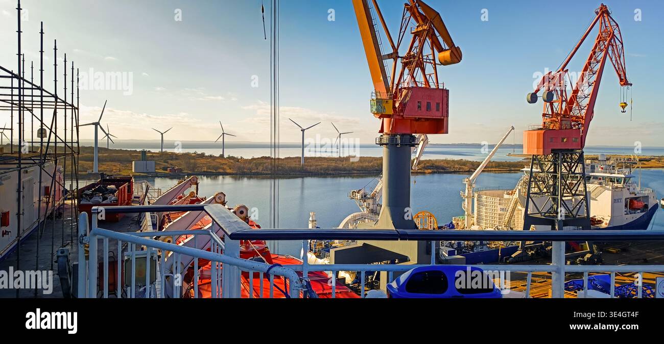 Vista da un ponte di navi presso le gru portuali e le turbine eoliche offshore in lontananza, scena industriale marittima con infrastrutture per energie rinnovabili. Foto Stock