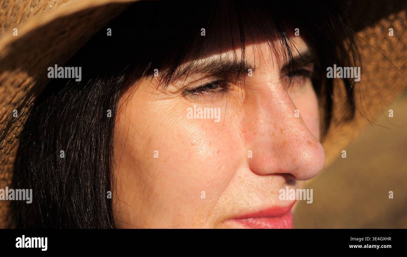 Agricoltore in cappello di paglia che guarda nel campo di grano e che esamina la piantagione al tramonto. Faccia ravvicinata di una bella donna adulta illuminata dalla luce Foto Stock