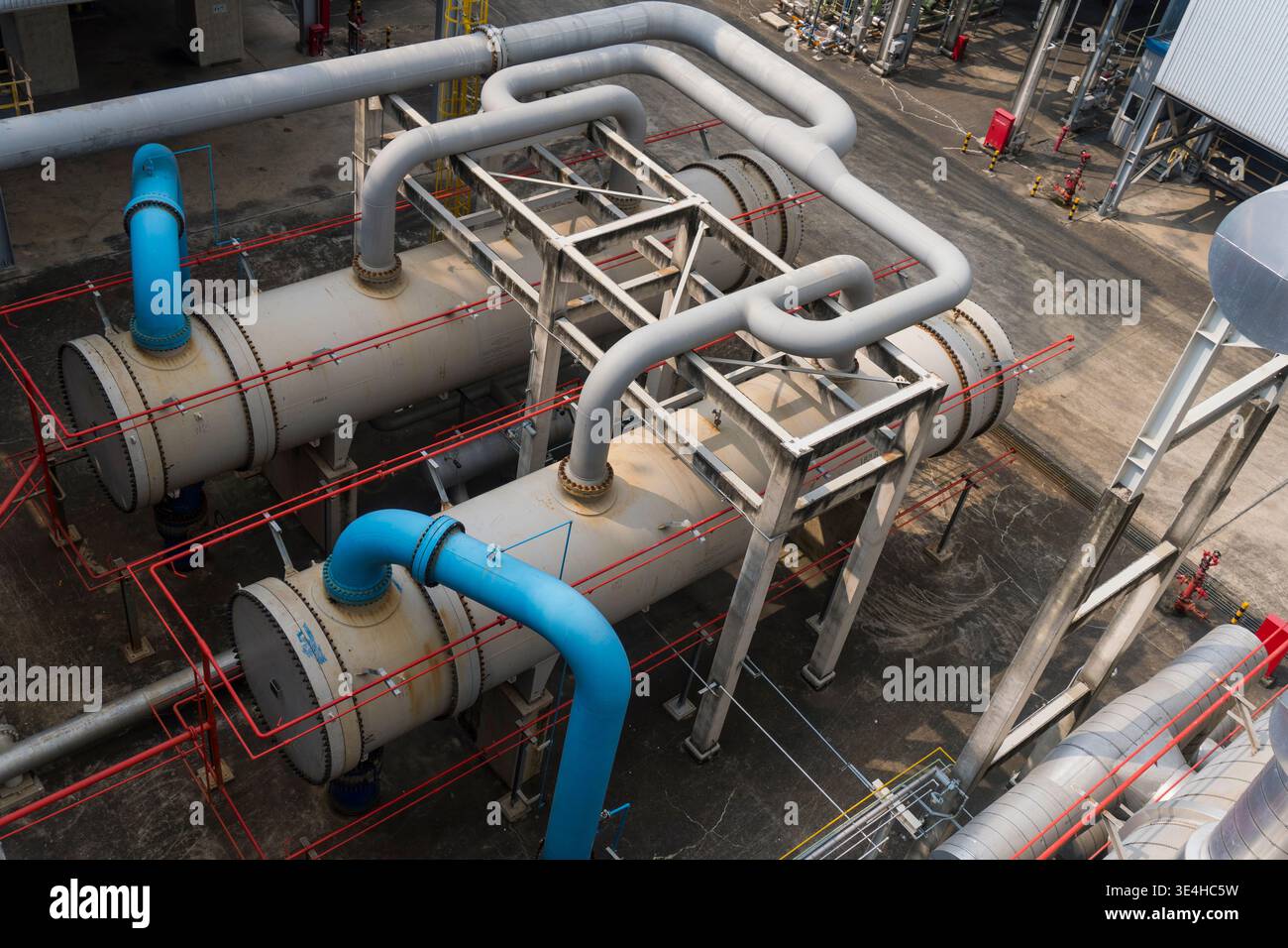 Tubi e serbatoi industriali in ambienti esterni. I tubi curvi e si collegano con i telai di supporto. I tubi blu e grigi contrastano con il terreno in calcestruzzo. Rosso sa Foto Stock