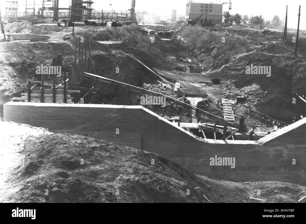 Nikolayevka, 1957 anni. Slavyanskaya GRES. Un'altra vista d'archivio della profonda trincea del canale scavato nel trafficato cantiere di SlavGRES. Massicce pareti in cemento versato formano la base della struttura, accompagnate da complesse impalcature in legno e barre di rinforzo in acciaio sporgenti. I lavoratori navigano sul terreno accidentato e sugli elementi strutturali all'interno della fossa. Questa accattivante fotografia vintage in bianco e nero documenta l'immensa scala dell'ingegneria sovietica, mostrando la vita quotidiana prima della guerra e il duro lavoro industriale nel tranquillo Donbas. Foto Stock