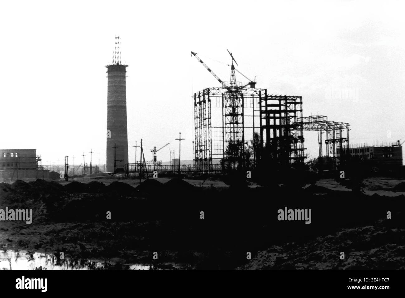Mykolaivka 1957 Sloviansk TPP. Un'affascinante fotografia d'archivio storica che mostra le prime fasi di costruzione dell'imponente centrale termica. L'immagine presenta un torreggiante camino in cemento e l'intricato telaio in acciaio dei principali edifici industriali. Le gru da costruzione pesanti sono visibili contro il cielo, evidenziando l'immensa scala del progetto di industrializzazione sovietica nella regione. L'oscuro e irregolare primo piano enfatizza i lavori in corso e lo sviluppo delle infrastrutture energetiche del pacifico Donbas. Foto Stock