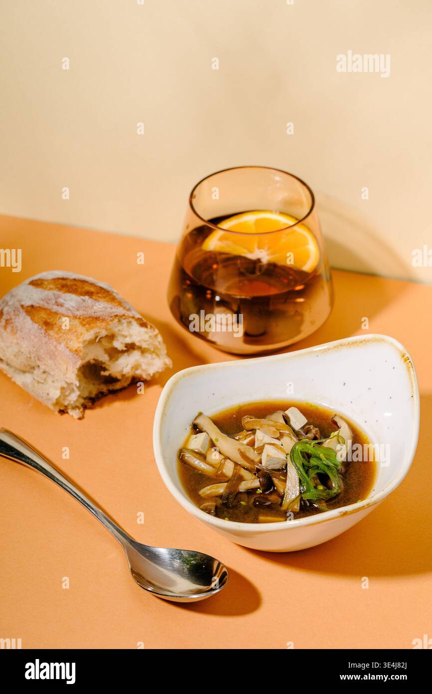 Zuppa di miso con cubetti di tofu, shiitake e funghi enoki in brodo dashi con alghe wakame e cipolla verde, servita in una ciotola accanto a una bevanda e pane Foto Stock