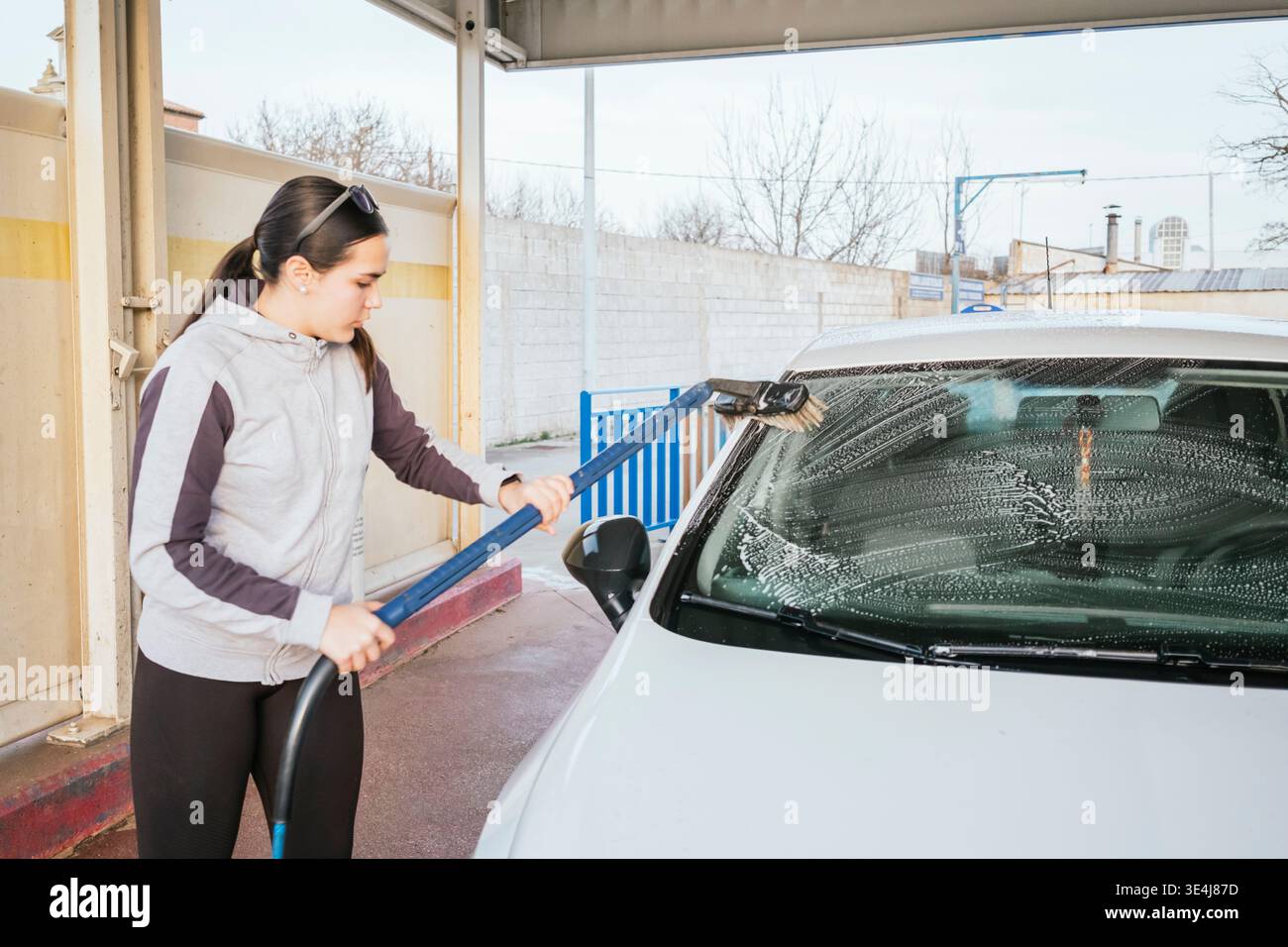 Adolescente che lava un'auto bianca in un autolavaggio self-service all'aperto, usando una lunga spazzola per strofinare il parabrezza con sapone durante il giorno. Foto Stock