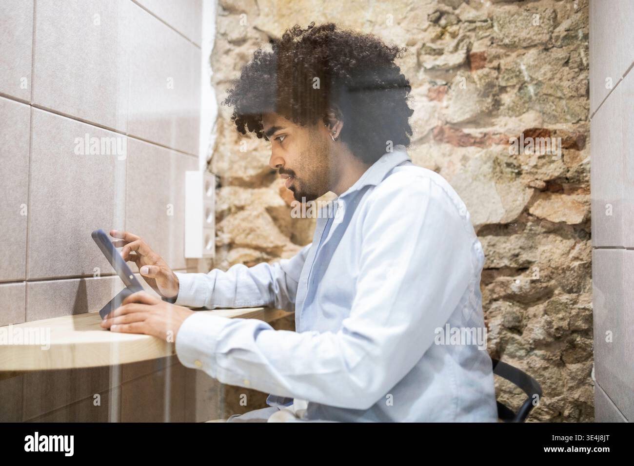 Vista laterale di un uomo latino che lavora in un ufficio moderno, utilizzando un tablet su una scrivania di legno. Professionista concentrato nella camicia da lavoro, contro la pietra rustica wal Foto Stock