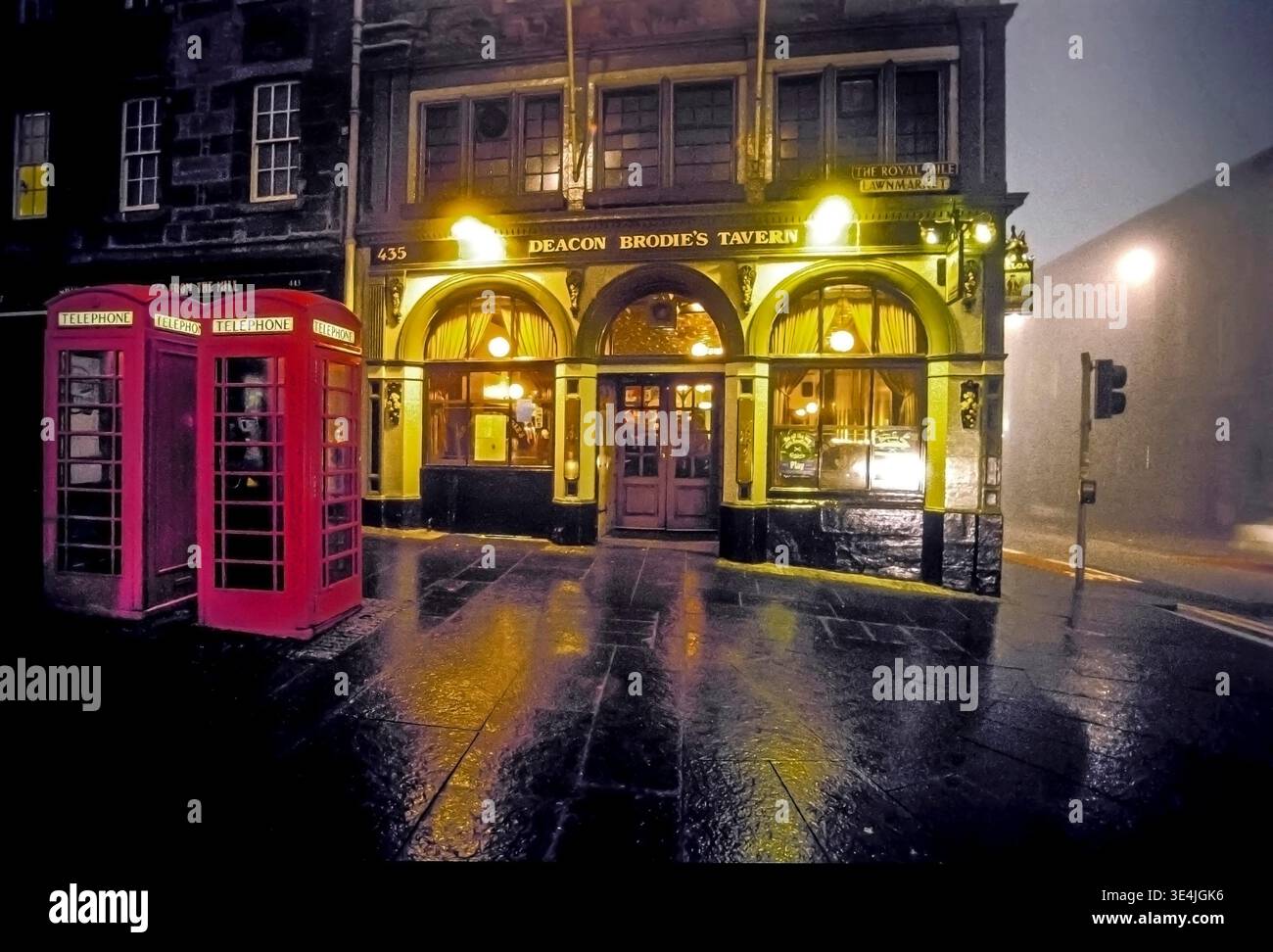 La calda luce della Deacon Brodie's Tavern si illumina di notte sul Royal Mile di Edimburgo, incorniciata da classiche cabine telefoniche rosse, ciottoli bagnati e riflessi nebbia Foto Stock