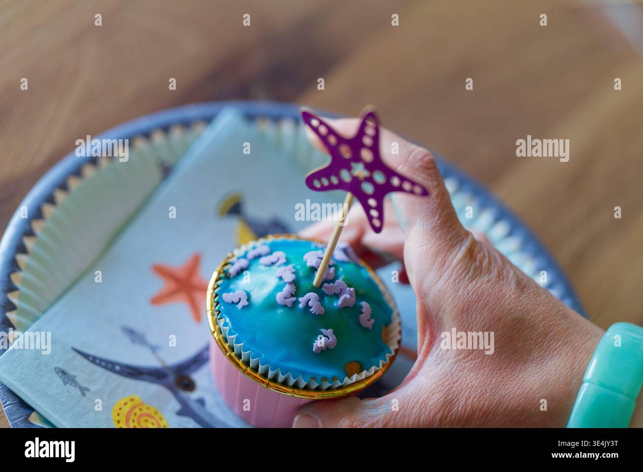 Una mano regge un cupcake decorato con glassa blu e piccoli spruzzi viola di creatura marina, sormontato da una decorazione viola con stelle marine su un tavolo di legno. Foto Stock