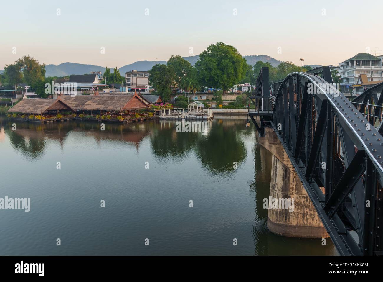 Kanchanaburi, Thailandia - 22 novembre 2024: Il ponte sul fiume Kwai attraversa il fiume Kanchanaburi con edifici lungo il lungomare. Foto Stock