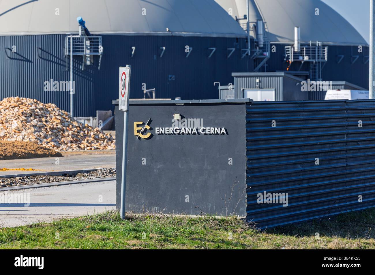 Cerna, Croazia - 27 febbraio 2026: Un cartello d'ingresso e un logo dell'impianto di produzione di biogas montati su una parete scura di un impianto industriale, con un grande serbatoio a cupola Foto Stock