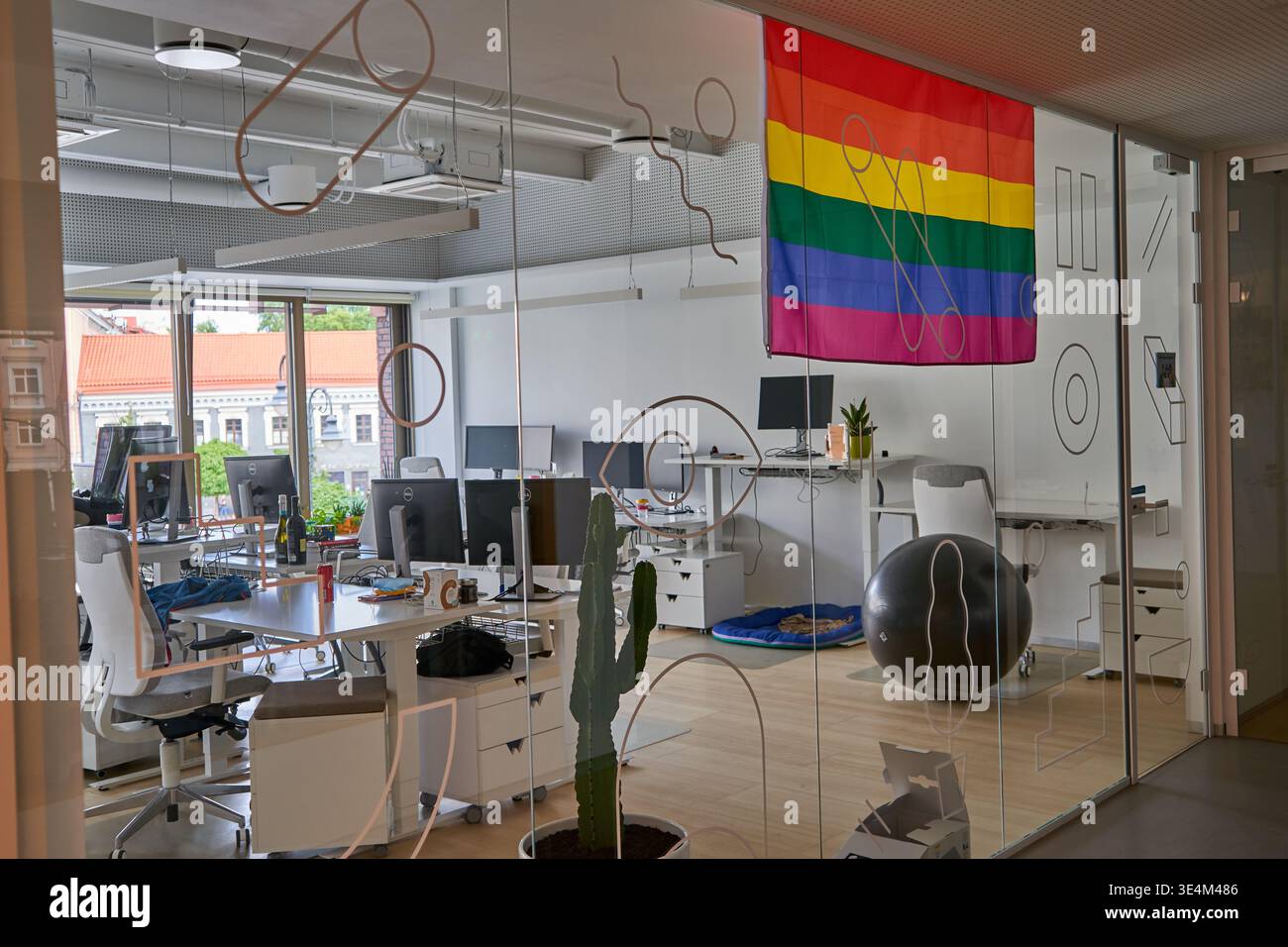 Un moderno spazio di lavoro da ufficio visto attraverso pareti di vetro decorate con arte astratta del filo metallico. Una bandiera arcobaleno LGBTQ+ Pride è appesa in modo prominente al soffitto Foto Stock