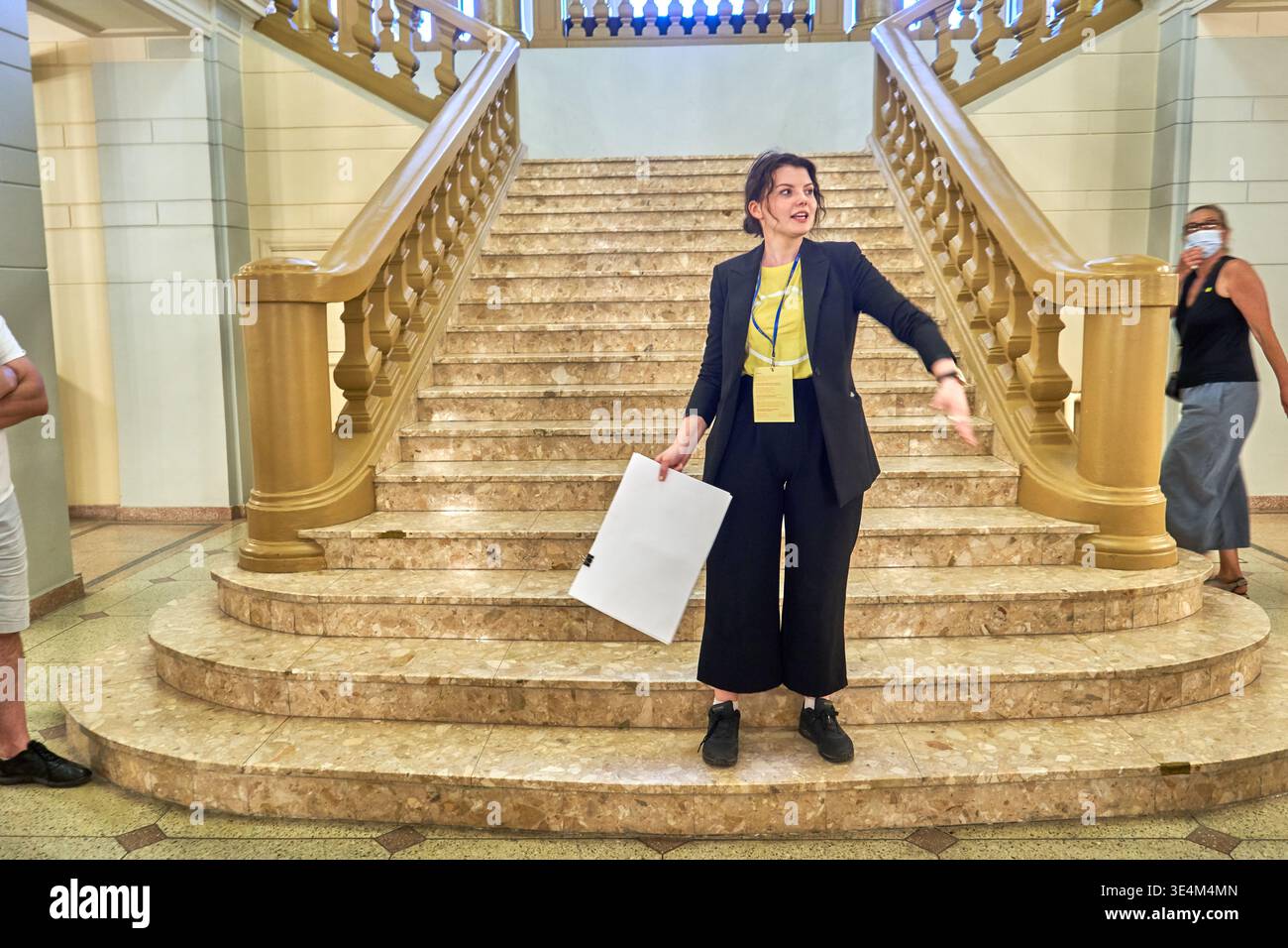 Una donna che indossa un blazer nero, pantaloni neri a gamba larga, scarpe nere e un top giallo si erge alla base di una grande scala di marmo, tenendo una Foto Stock
