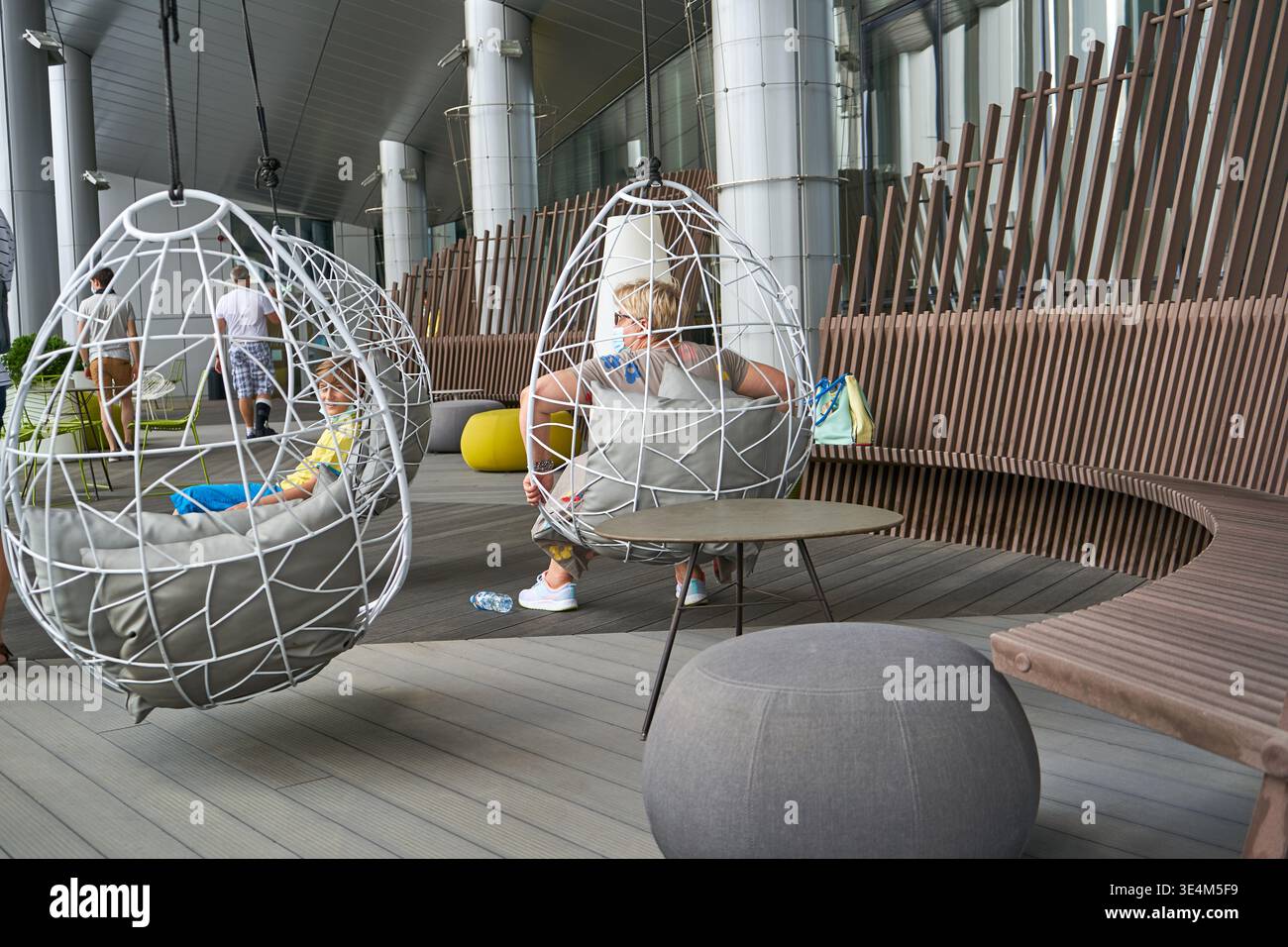 Due moderne sedie bianche sospese a forma di uovo con cuscini grigi sono sospese in una moderna area lounge interna-esterna. Un bambino in giallo Foto Stock