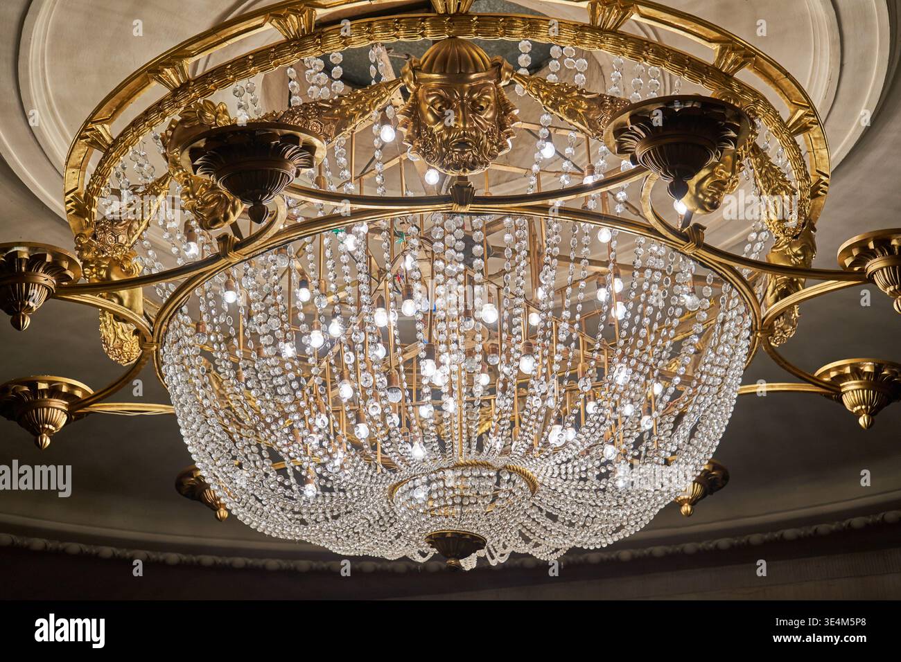 Vista ad angolo basso di un lampadario di cristallo classico decorato sospeso da un soffitto elaborato. L'impianto di illuminazione di lusso è caratterizzato da un'illuminazione complessa Foto Stock