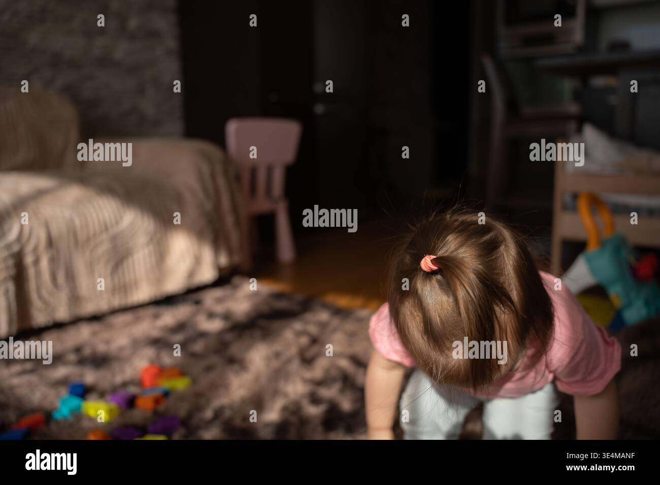 scena calda e candida di un bambino con una piccola coda di cavallo appoggiata in avanti su un tappeto di peluche in un soggiorno illuminato dal sole, blocchi colorati sparsi, sedia rosa e. Foto Stock
