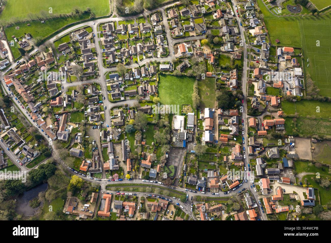 Vista aerea dall'alto verso il basso delle strade del villaggio di Bishop Monkton che ospitano giardini auto parcheggiate e un parco giochi scolastico che circondano campi verdi che formano un rur compatto Foto Stock