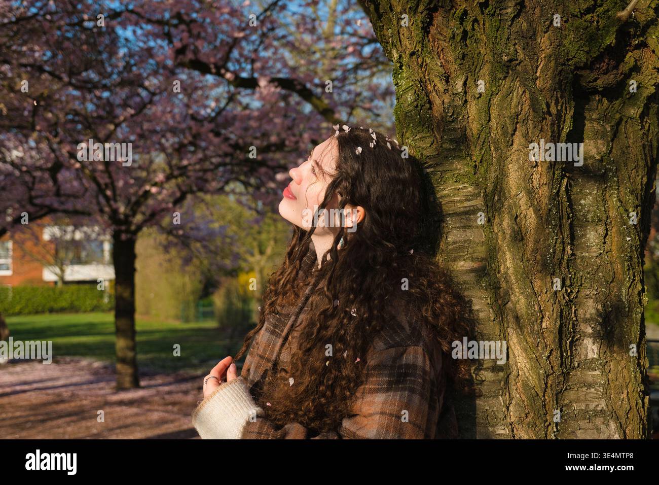 Giovane donna in piedi vicino a un albero sotto fiori di ciliegio in un parco. Luce solare calda, stagione primaverile e atmosfera tranquilla all'aperto. Foto Stock