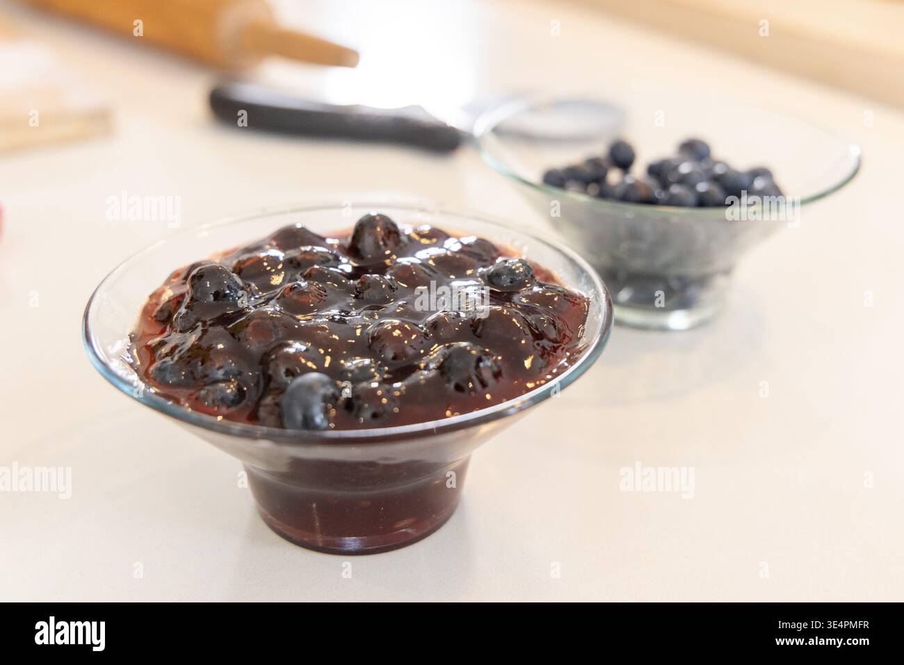 Ciotola in vetro di marmellata di mirtilli seduta sul bancone con mirtilli e mattarello sfocato Foto Stock