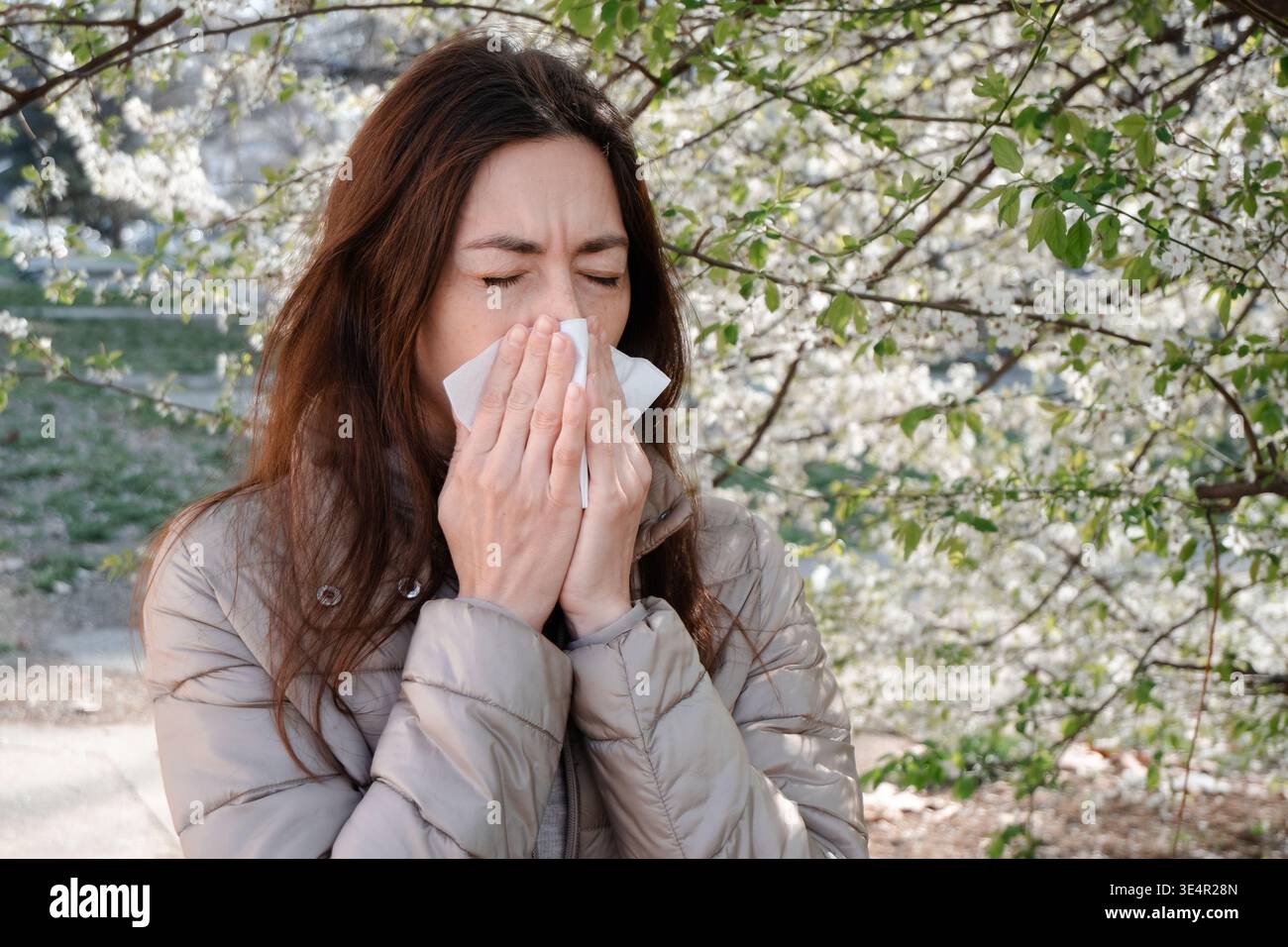 Donna che soffia il naso con un fazzoletto in un giardino primaverile fiorente. Sintomo di allergia stagionale, febbre da fieno disagio, problema di salute all'aperto. Foto Stock