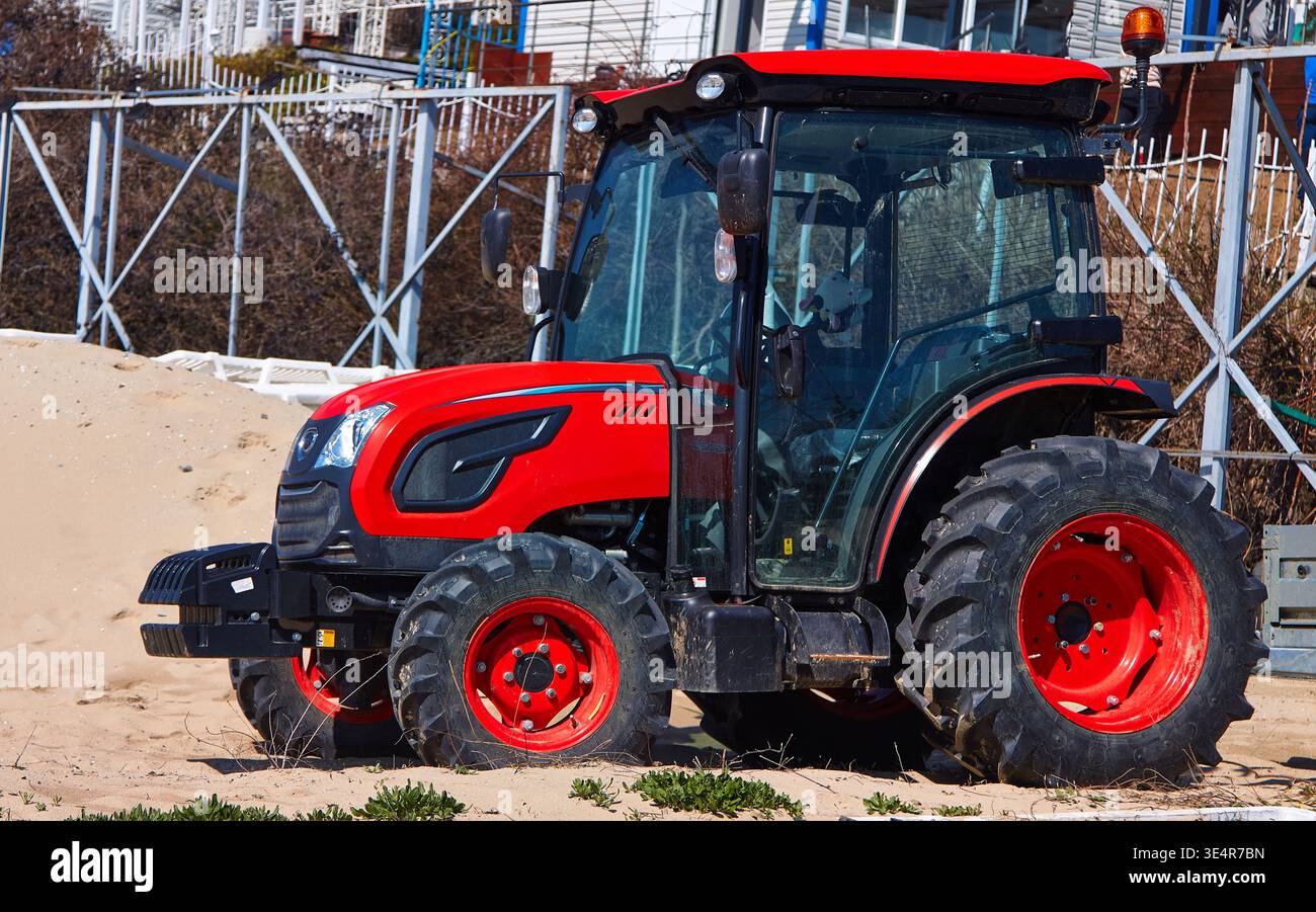 Trattore compatto in rosso e nero parcheggiato su un terreno sabbioso vicino a strutture costiere, con pneumatici di grandi dimensioni e cabina trasparente per una maggiore visibilità Foto Stock
