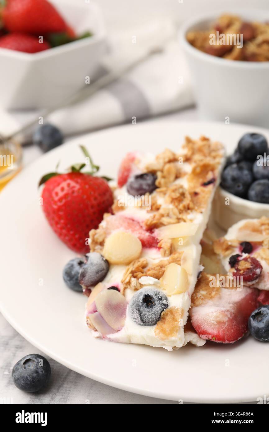Pezzi di gustosa corteccia di yogurt con frutti di bosco, scaglie di mandorle e muesli su tavolo di marmo bianco, primo piano Foto Stock