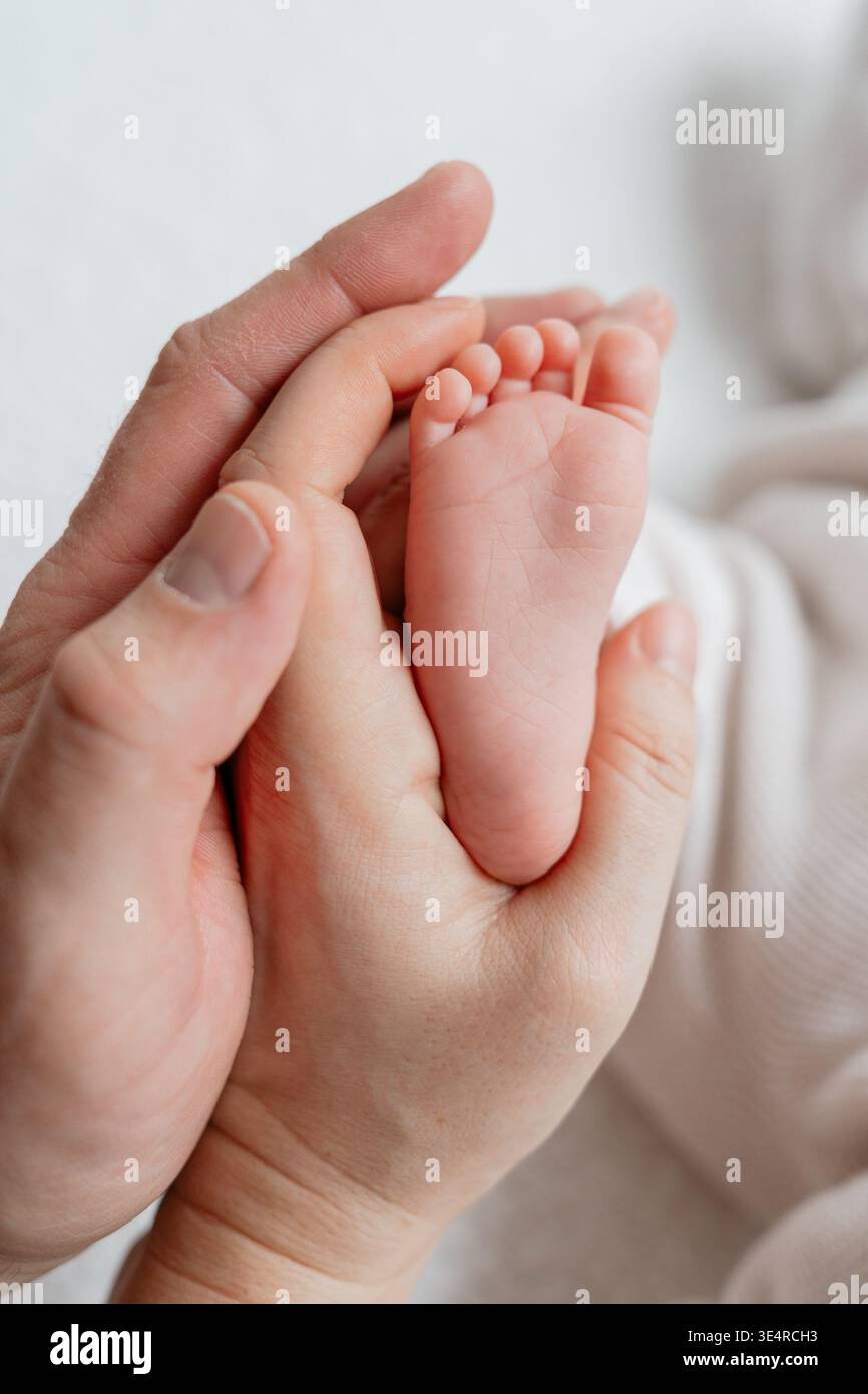 Primo piano verticale dei piedi del neonato, tenendoli delicatamente nelle mani dei genitori. L'illuminazione soffusa e la profondità di campo ridotta creano un'atmosfera intima ed emotiva. Foto Stock