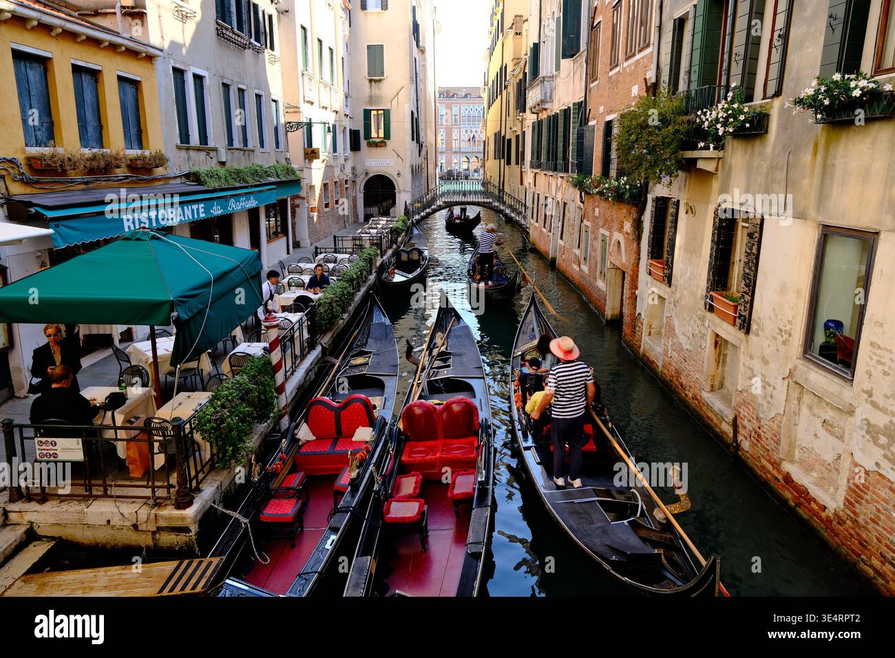 Venezia, Italia - 28 settembre 2025: Gondole in stretto canale veneziano, gondoliere in voga presso edifici storici e ristorante all'aperto sul lungomare. Foto Stock