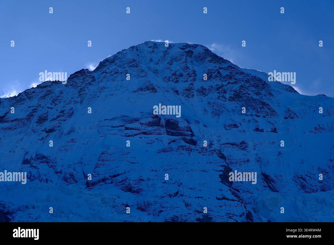 Primo piano della parete nord dell'Eiger durante l'ora blu, ripide superfici rocciose e spindrift che soffiano fuori dalla cresta della montagna Foto Stock