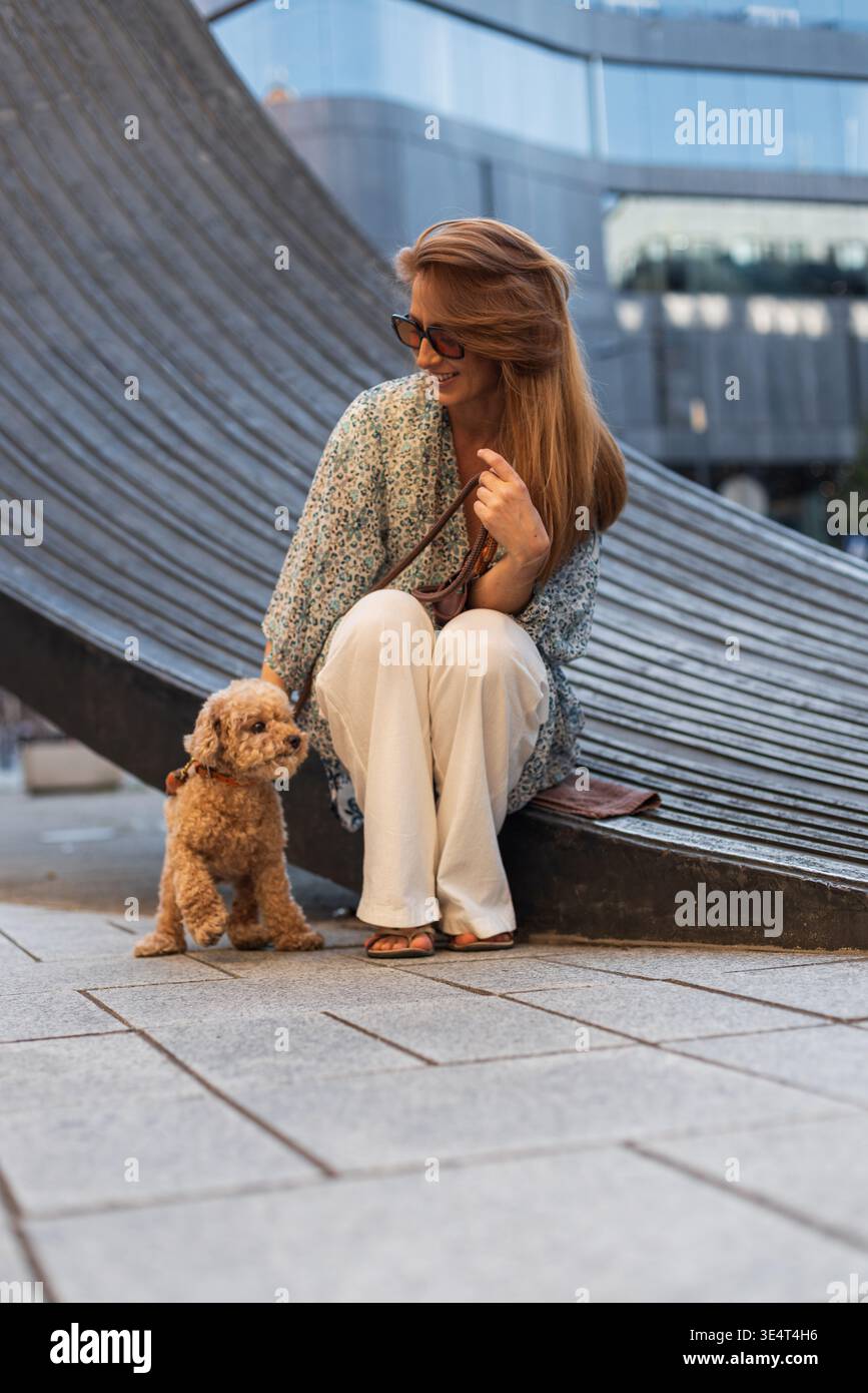 donna elegante seduta con il suo simpatico barboncino in una strada urbana. Il cane è al guinzaglio e entrambi si godono un momento di calma all'aperto, circondati Foto Stock