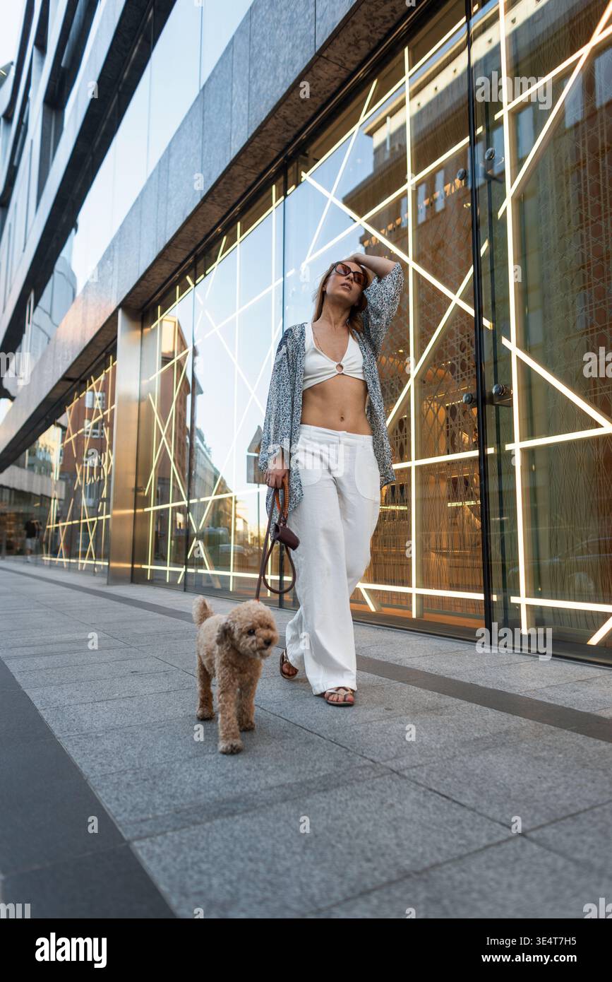 Elegante giovane donna con pantaloni bianchi e top di fascia che passeggia con il suo cane. Paesaggio urbano, moda di lusso e architettura moderna. Foto Stock