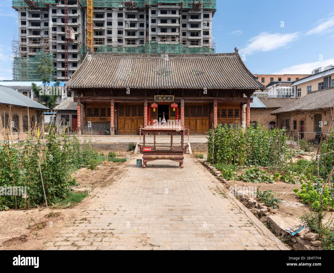 Un tradizionale cortile cinese caratterizzato da un sentiero pavimentato e orto, pesantemente oscurato da un massiccio e moderno cantiere edile. Il Foto Stock