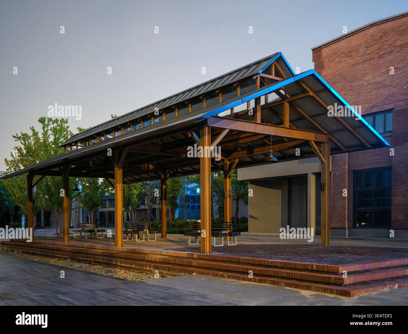 Un padiglione in legno del parco costruito su una piattaforma in mattoni rialzati, caratterizzato da sottili accenti di luce blu lungo il bordo del tetto scuro in una piazza vuota di notte. Il Foto Stock