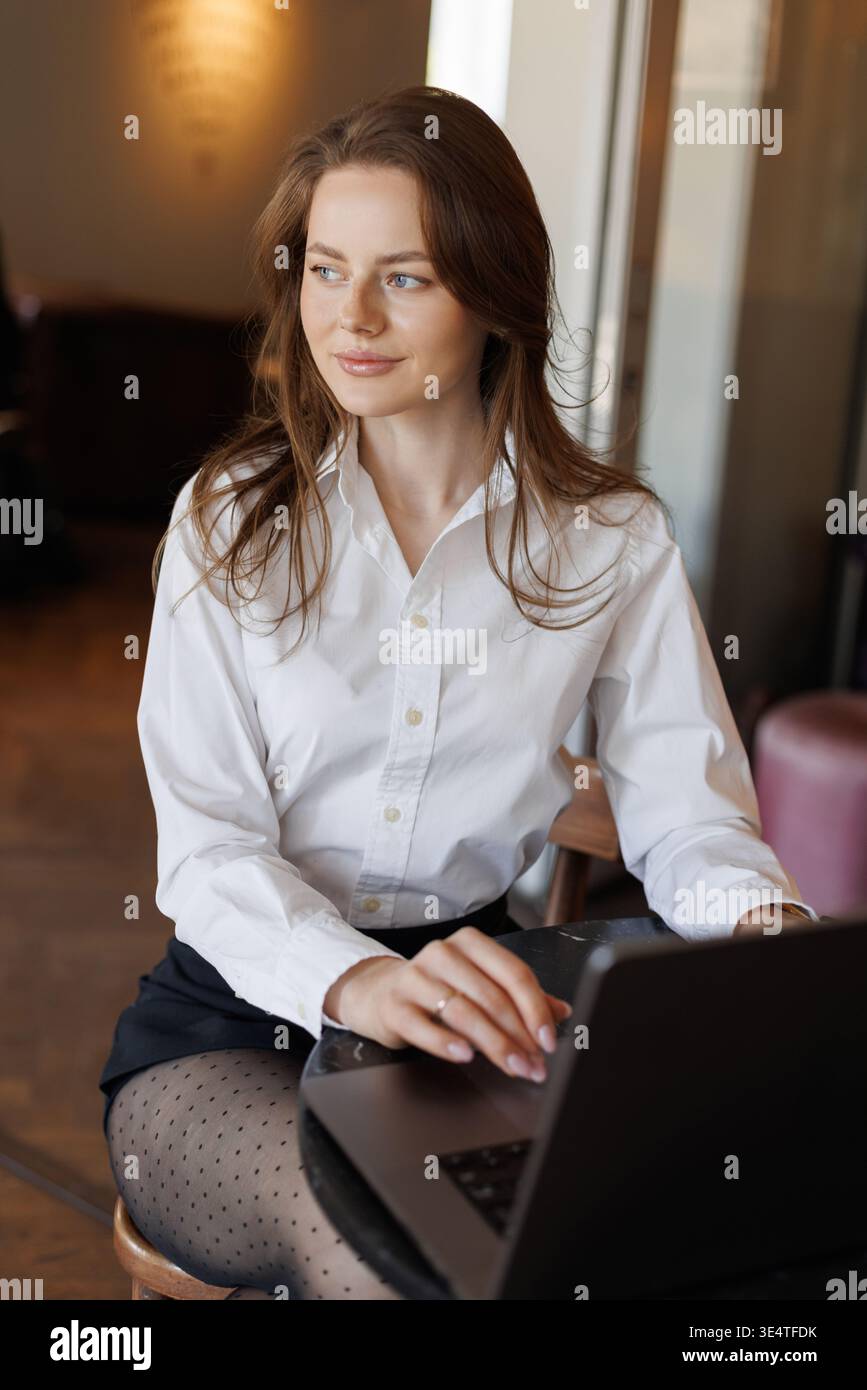Libero professionista al lavoro nell'accogliente caffetteria. Donna in abiti eleganti con laptop, circondata da moderni oggetti essenziali per lo spazio di lavoro come caffè, notebook e telefono. Foto Stock