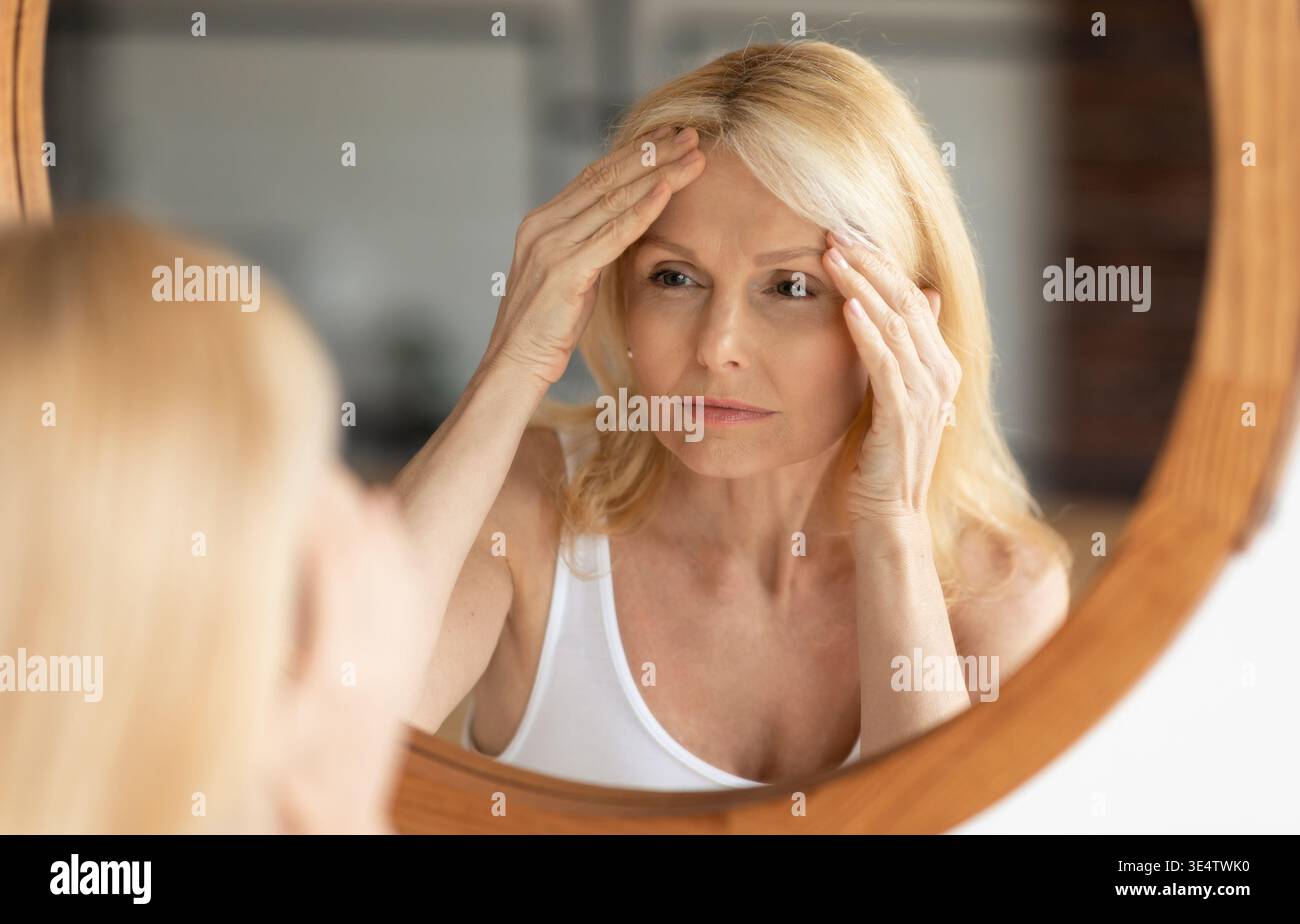 Donna anziana che guarda lo specchio e tocca la fronte, donna anziana sconvolta che esamina le linee sottili sul suo viso Foto Stock