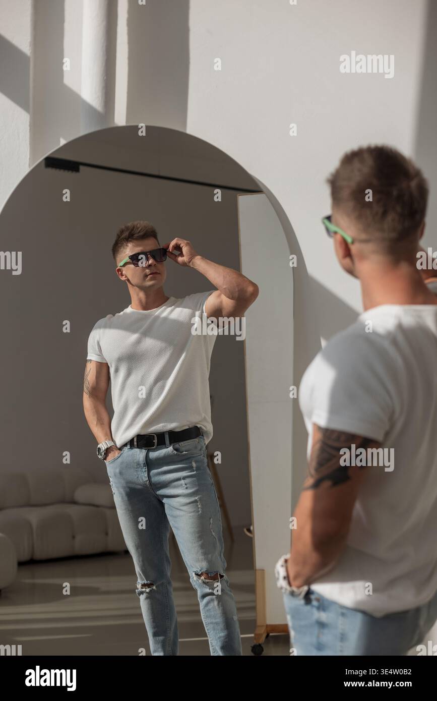 Uomo tatuato in t-shirt bianca e jeans che guarda il suo riflesso in uno specchio. Interni luminosi e moderni, luce solare che proietta ombre. Sicuro ed elegante Foto Stock