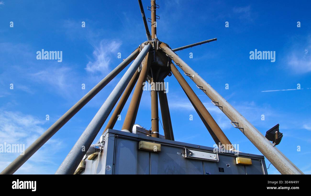 Un'antenna metallica si innalza contro un cielo blu, circondata da apparecchiature tecniche, che rappresentano l'infrastruttura di comunicazione in un ambiente industriale. Foto Stock