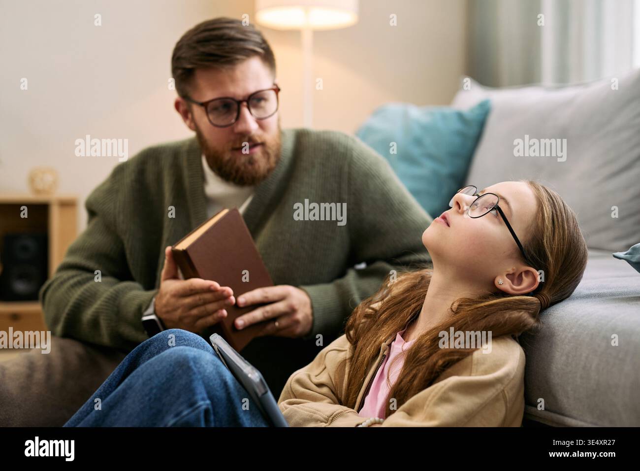 Uomo di mezza età caucasico in possesso di un libro che parla con una preadolescente caucasica che indossa gli occhiali seduti sul pavimento appoggiati sul divano ascoltando con un'espressione premurosa Foto Stock
