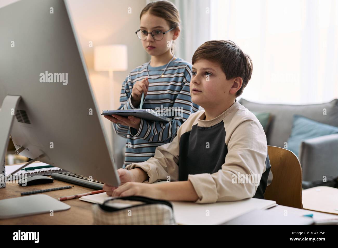 Ragazzo caucasico seduto alla scrivania utilizzando un computer desktop, mentre una ragazza caucasica in piedi accanto a un tablet digitale, entrambi i bambini si concentrano sullo schermo in un ambiente domestico Foto Stock