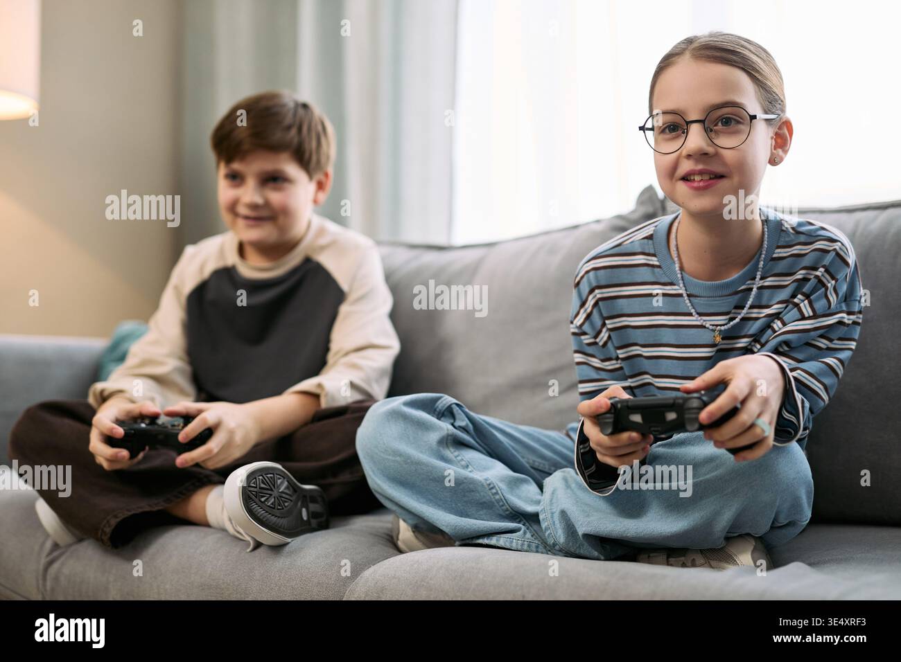 Due bambini caucasici seduti sul divano che giocano a videogiochi con controller, ragazzi e ragazze che si concentrano sullo schermo, entrambi apparendo impegnati e sorridenti, ambiente domestico informale, postura rilassata Foto Stock