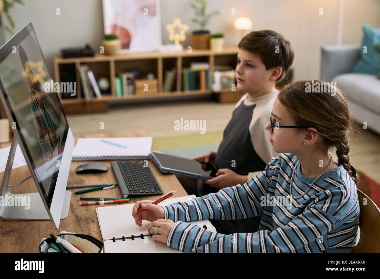 Ragazza caucasica e ragazzo caucasico seduti alla scrivania utilizzando un computer desktop per studiare, ragazza che scrive su un notebook mentre un ragazzo tiene un tablet digitale, entrambi concentrati sullo schermo Foto Stock