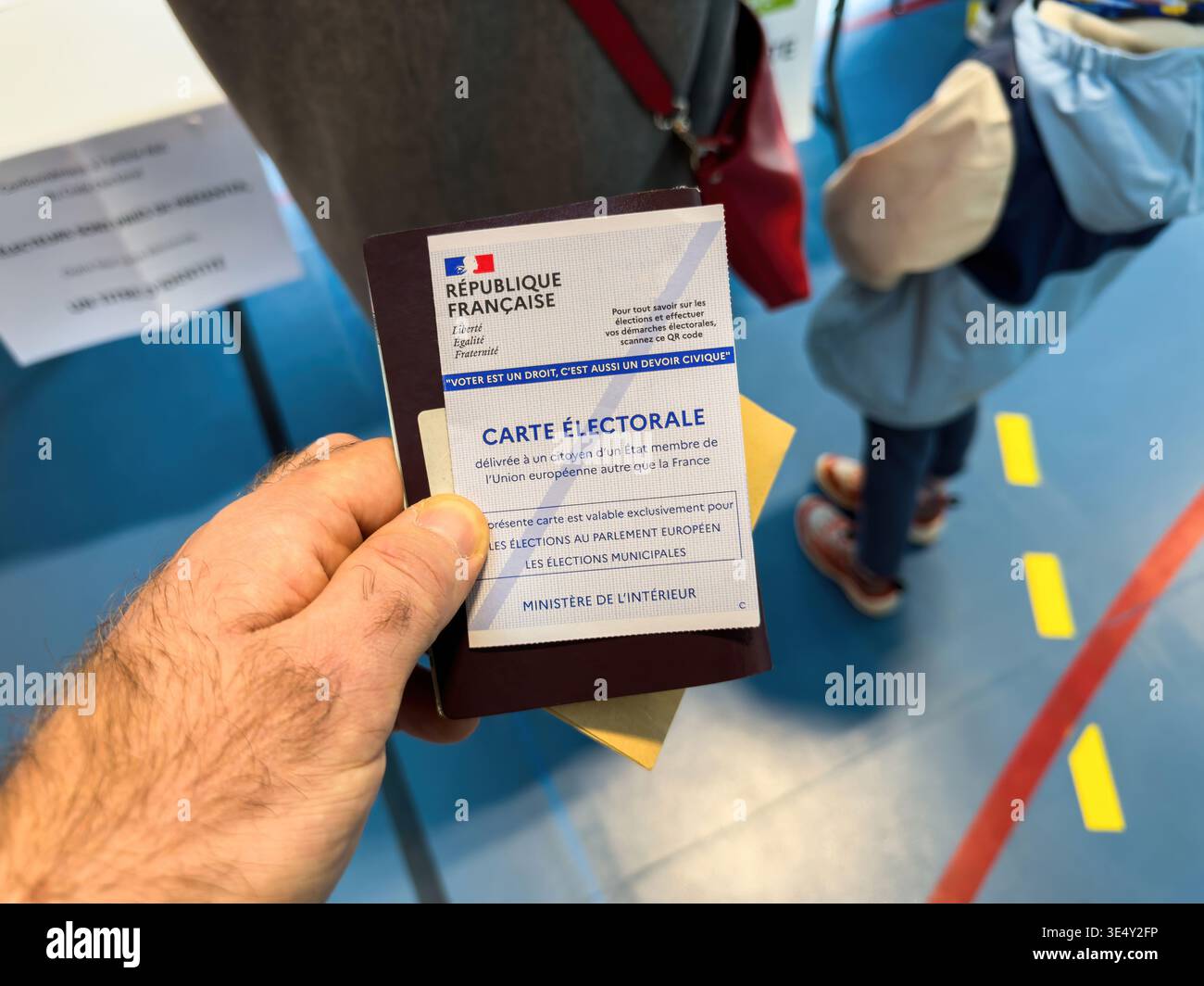 Parigi, Francia - 22 marzo 2026: Mano che tiene in mano il cartellino elettorale francese al seggio elettorale durante le elezioni comunali con gli elettori in coda e con la partecipazione civica Foto Stock